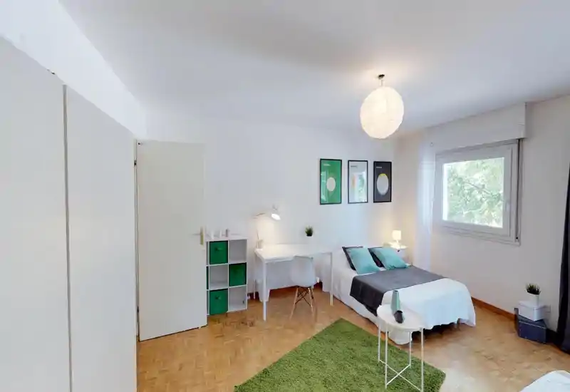 Bright, well-staged bedroom with a double bed, desk and natural light from a window. Clean parquet floor, green accents and simple modern decor create a calm, inviting space.