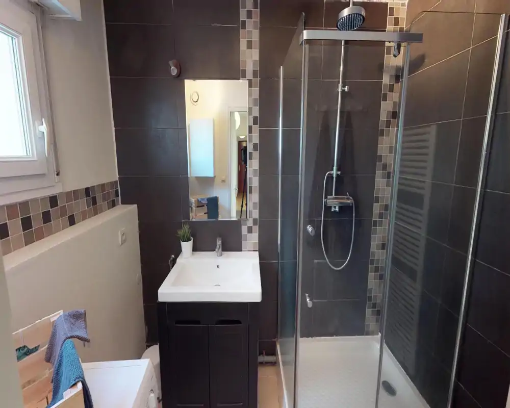 Modern tiled bathroom with a glass shower enclosure, wall-mounted rain shower, vanity with sink and a small mirror; neutral dark tiles and mosaic accents provide a contemporary look.