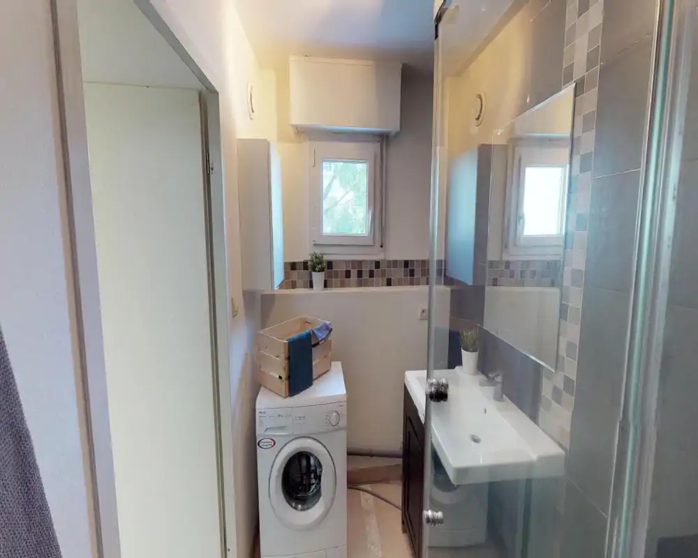Compact bathroom with a sink, mirror, shower screen and a washing machine beneath a small window. Neutral tiles and a tidy layout create a functional, well-lit space ideal for practical use.