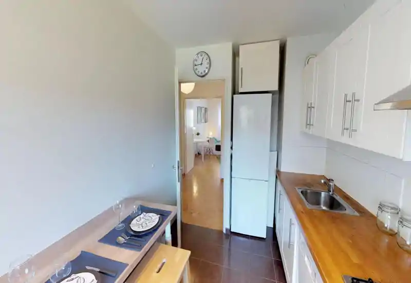 Cuisine compacte et lumineuse avec placards blancs, plan de travail en bois, évier en inox et réfrigérateur toute hauteur ; une petite table est visible à gauche.