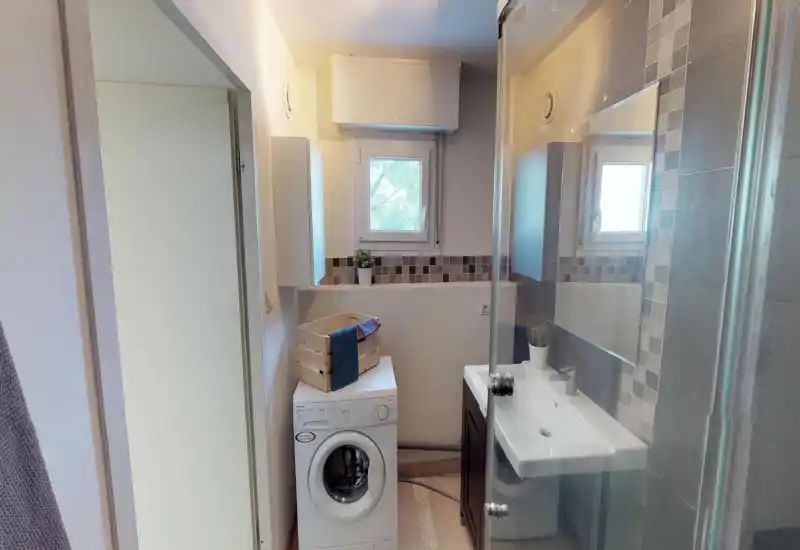 Salle de bains compacte avec lavabo, miroir, paroi de douche et machine à laver sous une petite fenêtre. Carrelage neutre et agencement soigné pour un espace fonctionnel et bien éclairé.