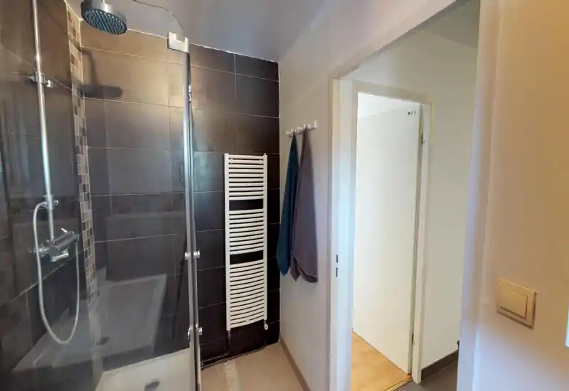 Salle de bain moderne carrelée avec douche vitrée, pommeau pluie et sèche-serviettes; tons neutres et bonne luminosité.