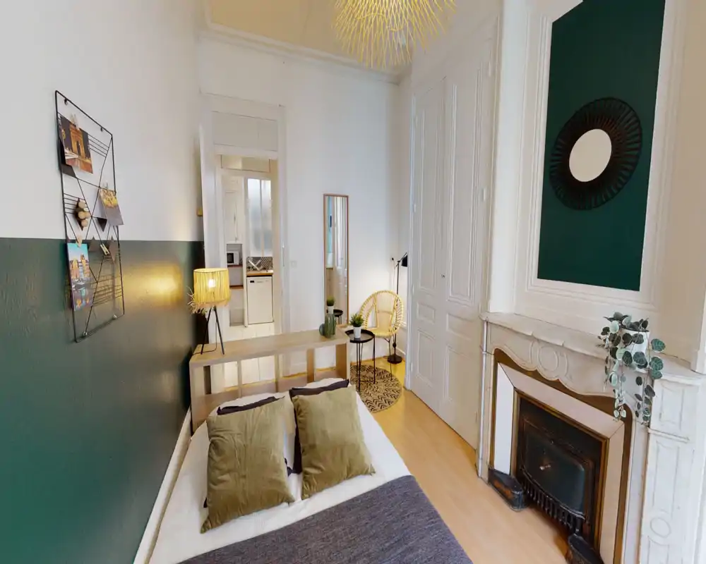 Chambre lumineuse et élégante avec un grand lit confortable, coussins texturés et décorations vertes. La pièce offre des moulures d'origine, une cheminée décorative et un coin salon près de la penderie.