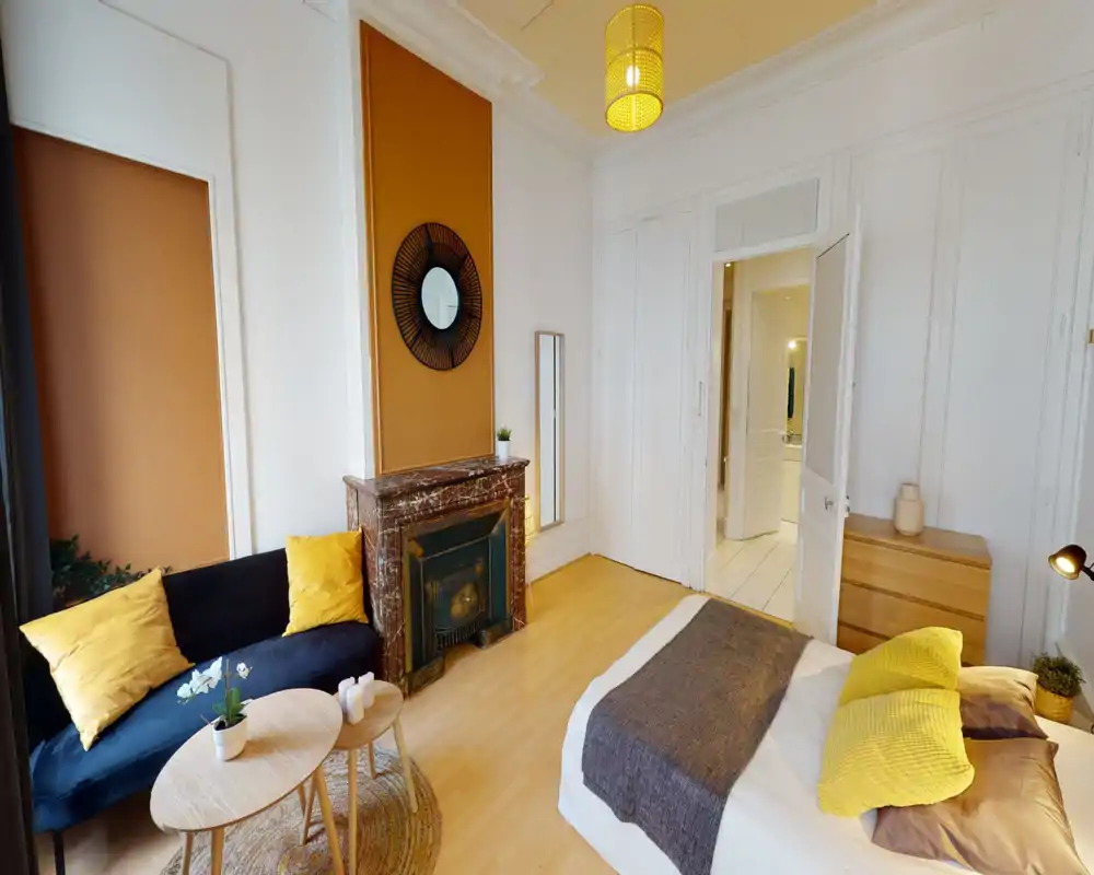Chambre lumineuse et élégante avec lit double habillé de linge neutre et coussins jaunes, une cheminée décorative et un petit coin salon avec canapé bleu marine et tables basses en bois.
