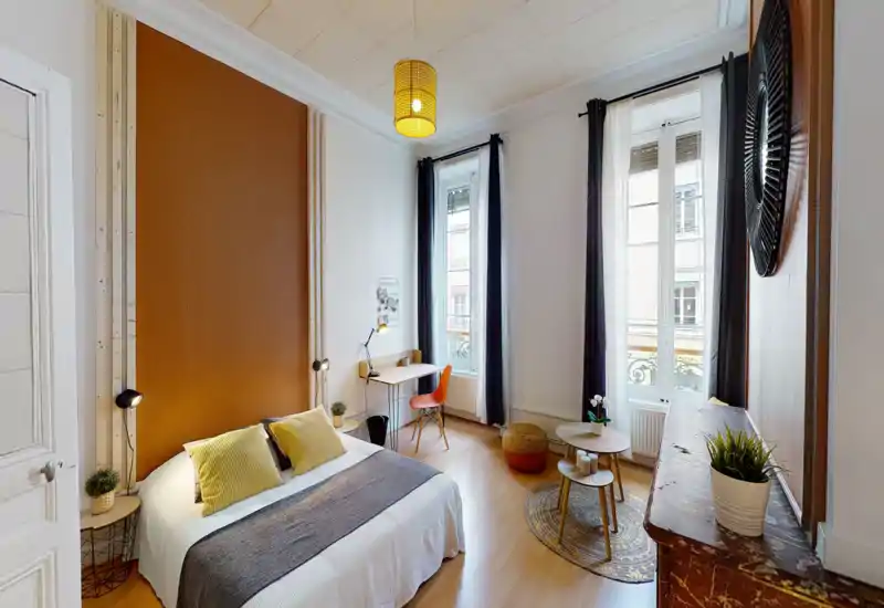 Bright, well-styled bedroom with a comfortable double bed, warm accent wall and large windows providing abundant natural light. A small desk and seating area make it suitable for both rest and work.