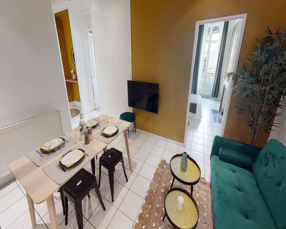 Espace séjour lumineux et stylé avec canapé vert en velours, tables gigognes, TV murale et table dressée — idéal pour des séjours détendus et petits dîners.