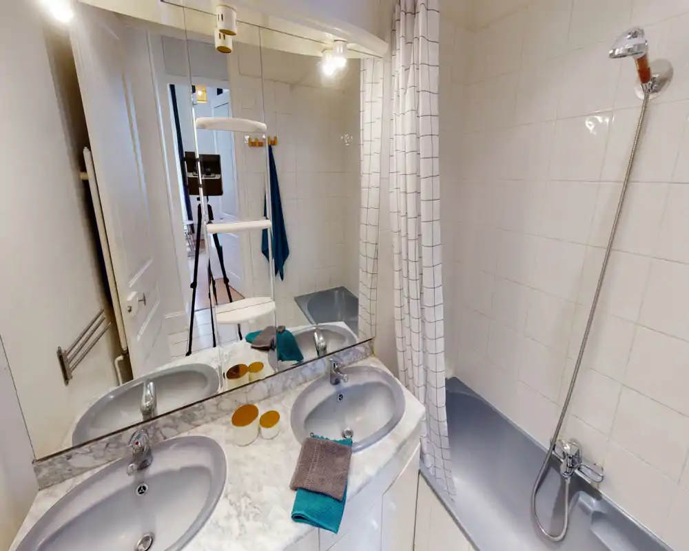 Salle de bains propre et bien éclairée avec baignoire, douchette, double vasque sur plan en marbre et grand miroir. Carrelage neutre et rideau de douche à carreaux pour une impression nette et soignée.