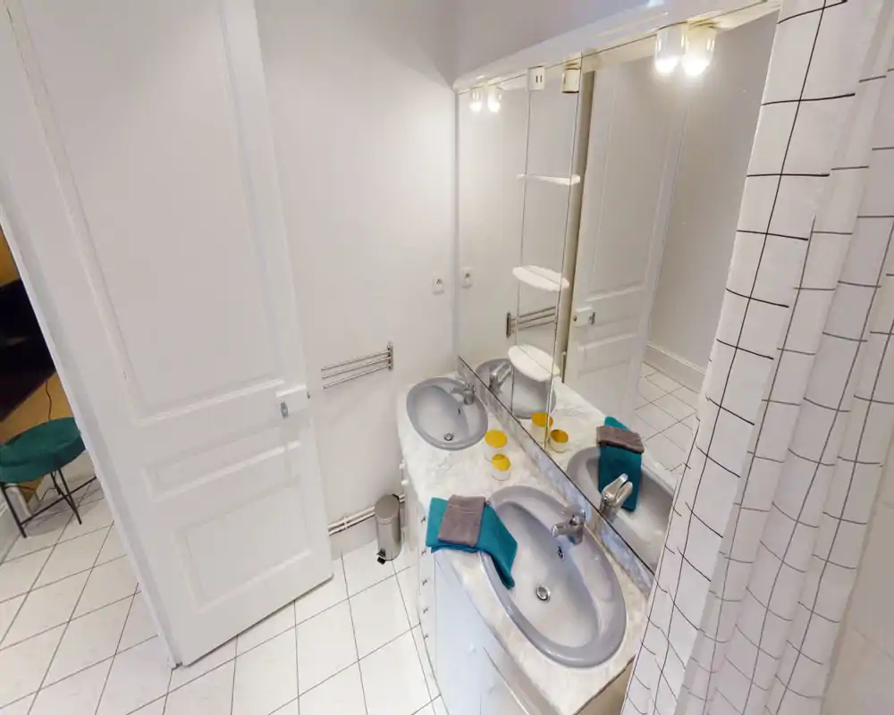 Salle de bains propre et lumineuse avec double vasque, grand miroir et sol carrelé ; rideau de douche visible à droite.
