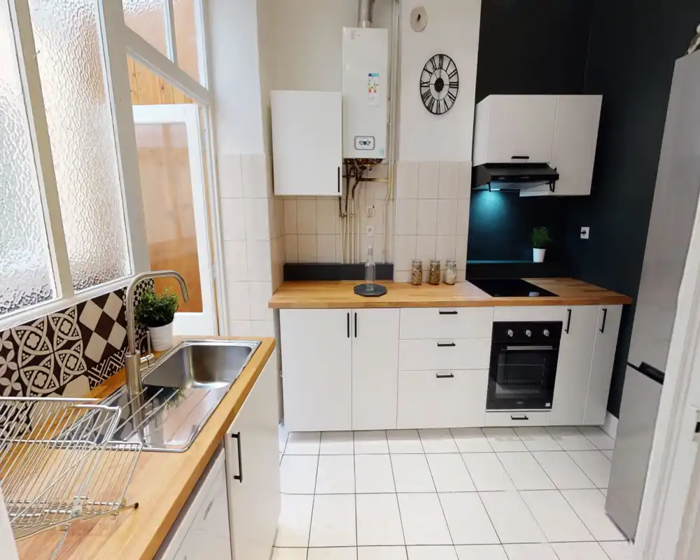 Cuisine moderne et compacte avec plans de travail en bois, meubles blancs, four encastré, plaque de cuisson et évier en inox près d'une fenêtre dépoli — lumineuse et bien mise en scène pour une annonce.