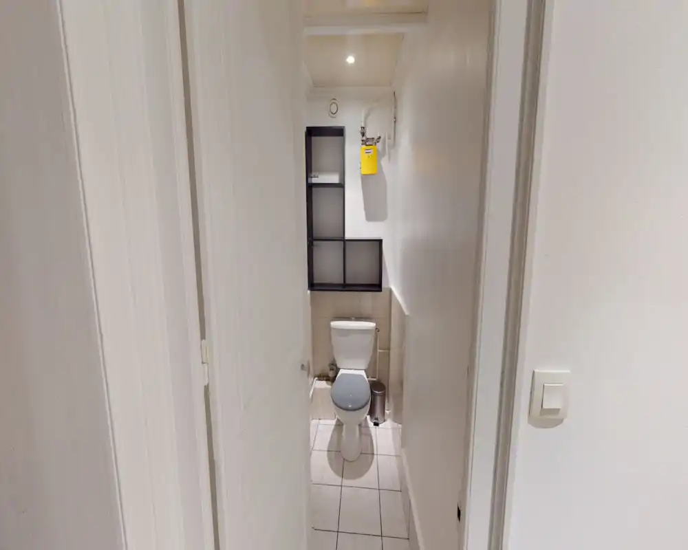 Petite salle de bain avec WC, sol carrelé et petite étagère murale au-dessus de la chasse d'eau. Murs blancs propres et éclairage doux offrant un espace fonctionnel et neutre.