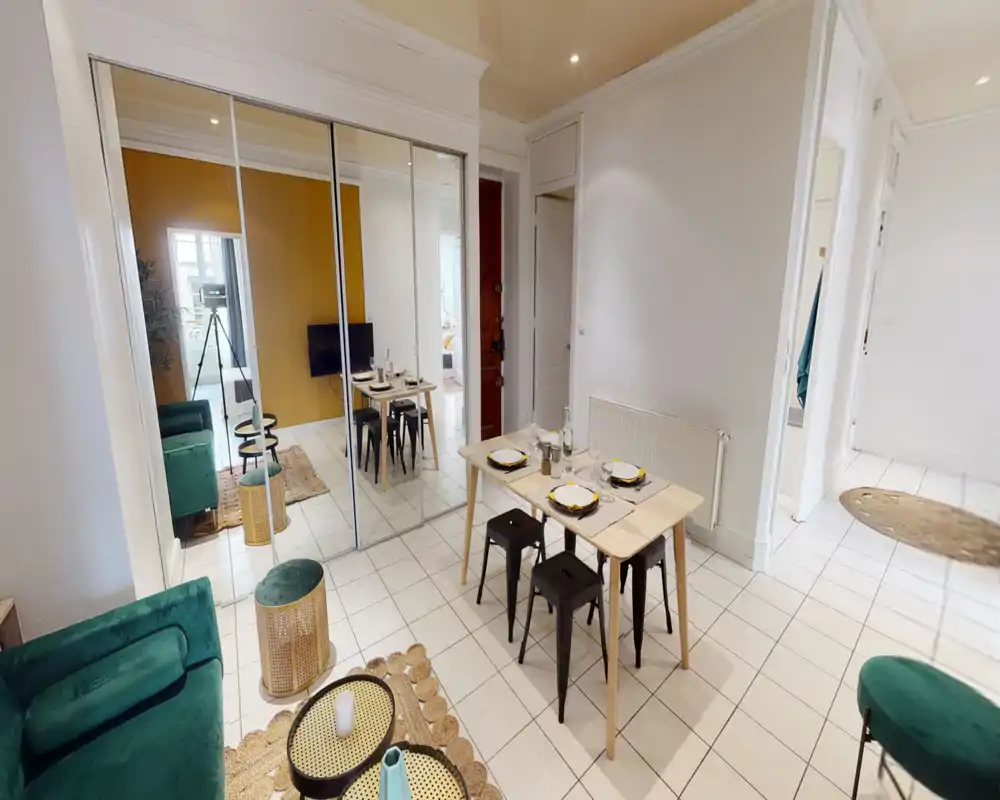 Espace de vie lumineux et aménagé avec une petite table à manger et un canapé vert ; des placards à portes miroir agrandissent la pièce et un mur moutarde apporte de la couleur.