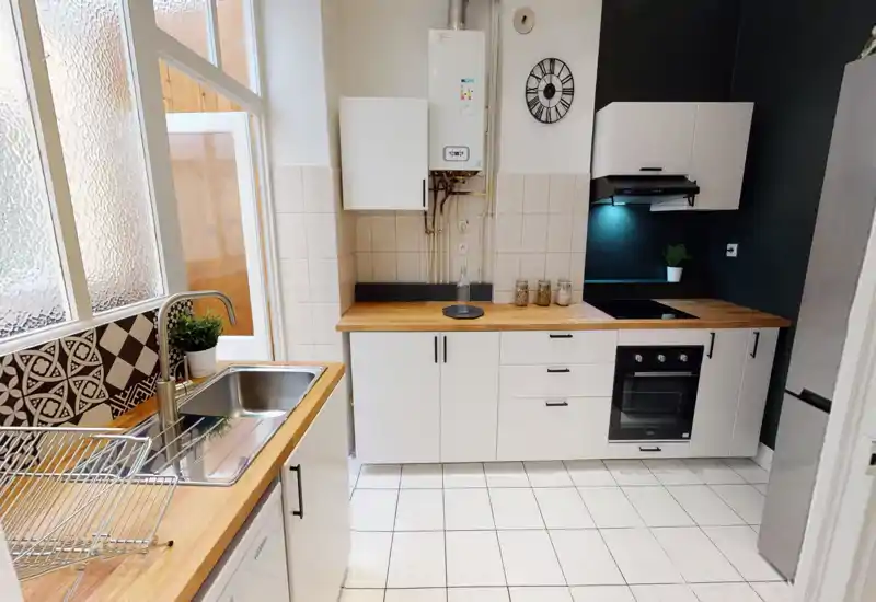 Modern, compact kitchen with wooden countertops, white cabinetry, built-in oven and cooktop, and a stainless-steel sink by a frosted window — bright and neatly styled for rental photos.