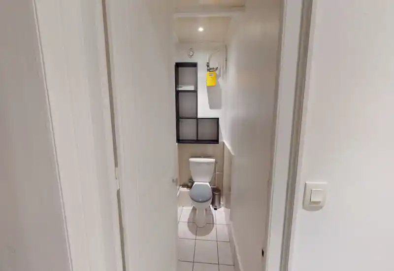 Compact bathroom with a toilet, tiled floor and a small wall shelf above the cistern. Clean white walls and soft overhead lighting provide a neutral, functional space.