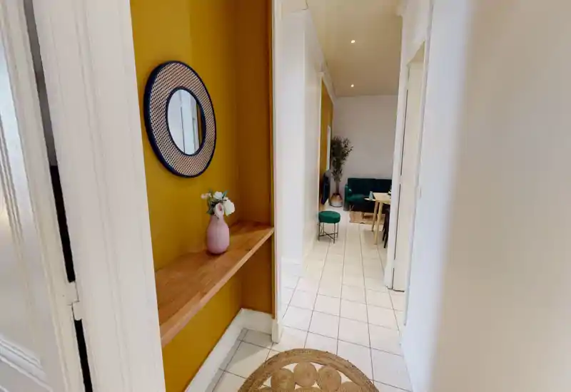 Entrance corridor leading into a living area with a green sofa, small stool and dining table visible in the distance; decorative yellow inset with mirror and vase adds style.