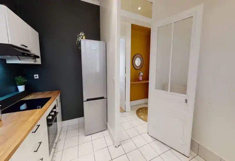 Modern compact kitchen with white cabinets, wood countertop, built-in oven and a stainless refrigerator. Clean tiled floor and tasteful contrast wall create a neat, bright cooking space.