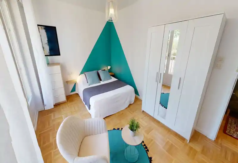 Camera da letto luminosa e ben arredata con letto matrimoniale davanti a una parete accentuata triangolare turchese, mensoline e lampade accanto al letto, armadio bianco con specchio e un angolo relax accogliente.