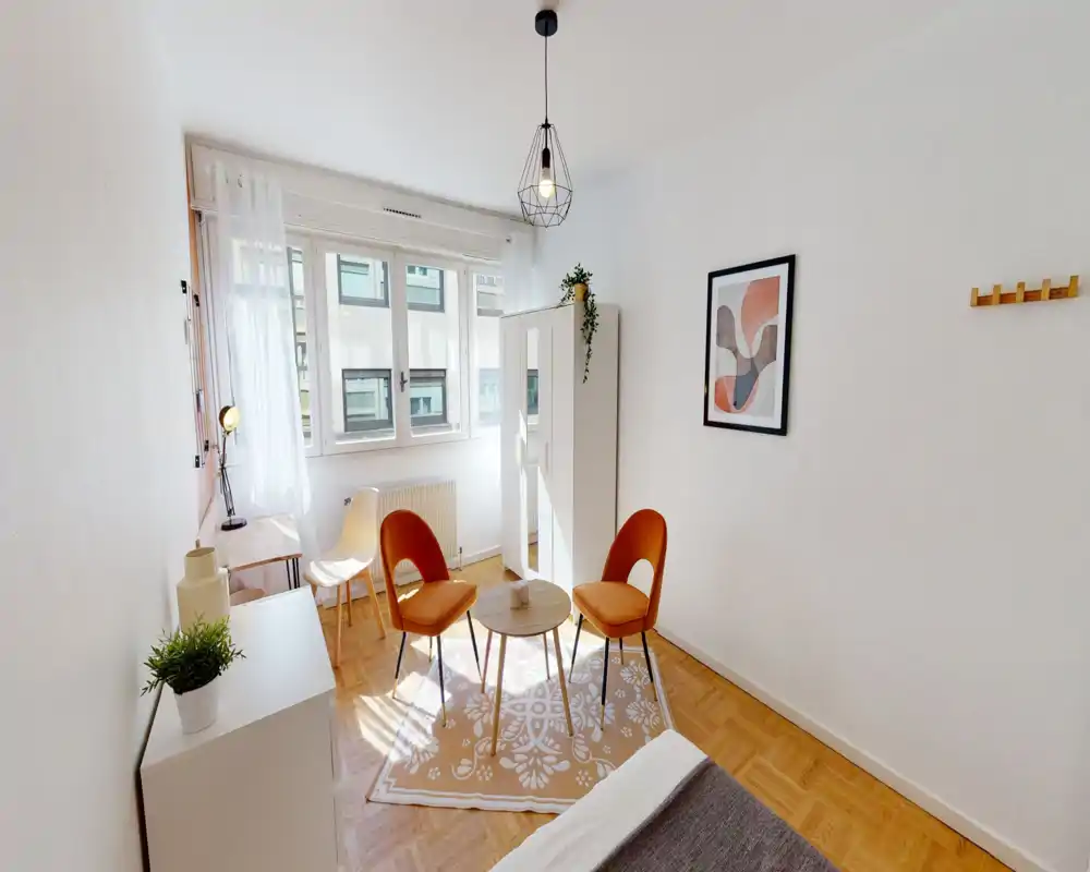 Bright, well-staged bedroom corner with a small seating area, wardrobe and natural light from a large window — ideal for single or short-term stays.