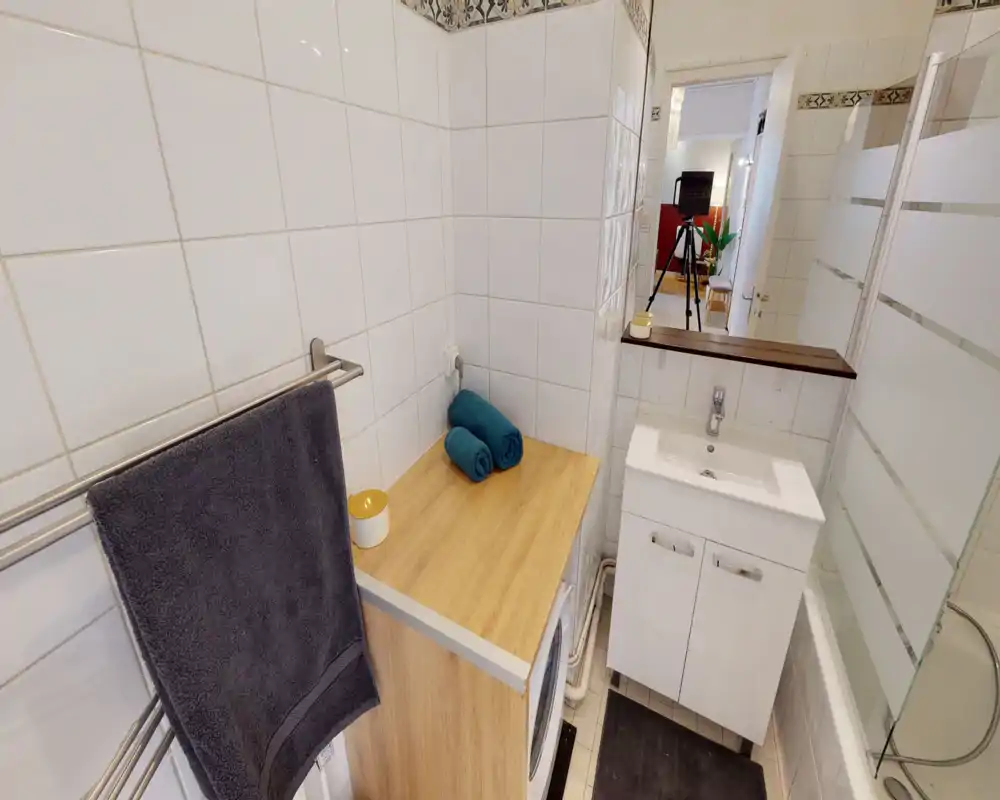 Bagno compatto e ordinato con pareti piastrellate bianche, lavabo con mobile, vasca con pannello di vetro satinato, portasciugamani e lavatrice sotto un piano in legno. Spazioso e funzionale per un appartamento cittadino.