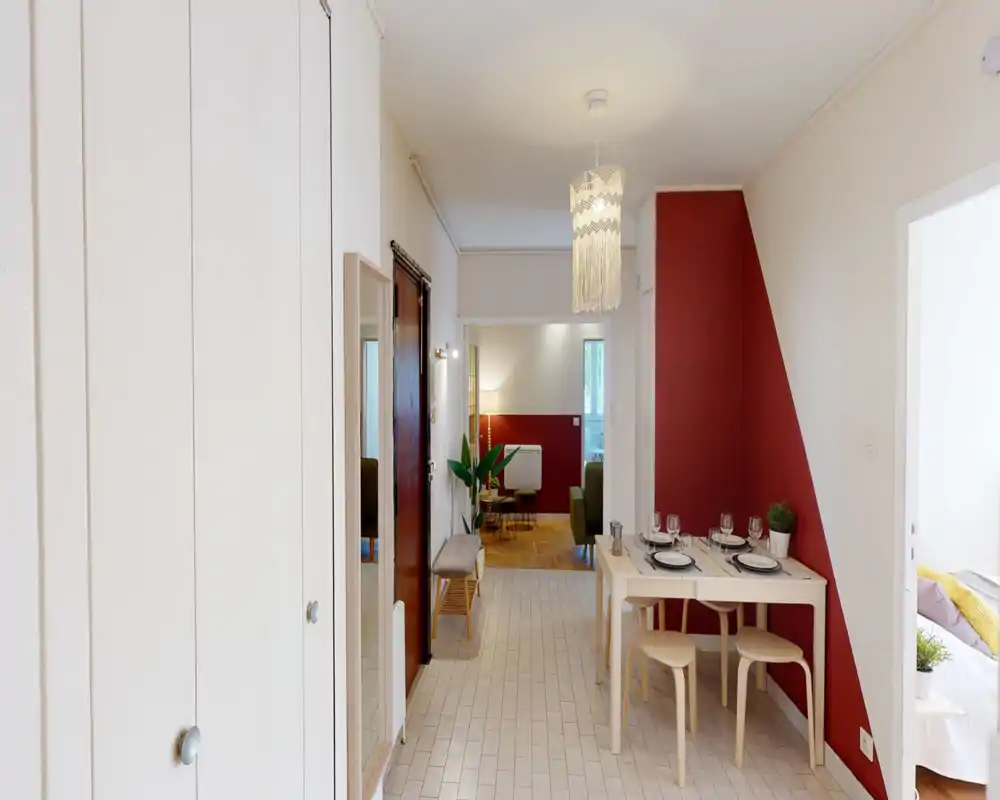 Zona giorno luminosa e ben allestita vista dall’ingresso, con un piccolo tavolo da pranzo, una parete rossa di accento e la vista su un soggiorno accogliente con luce calda.