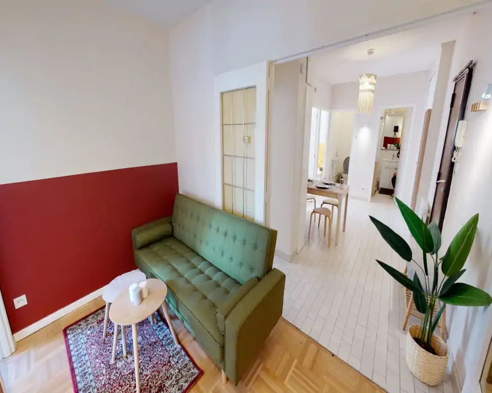 Zona giorno luminosa e curata con un divano verde capitonné, piccoli tavolini e un tappeto decorato; l'ambiente aperto si collega alla zona pranzo e all'ingresso.