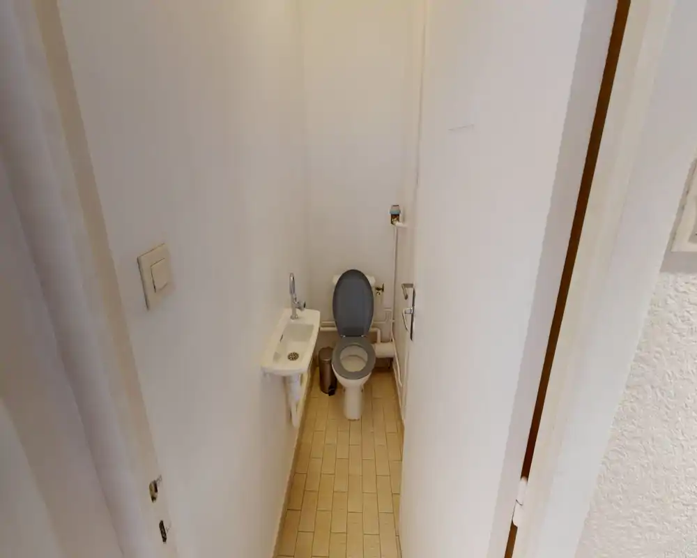 Bagno compatto e ordinato con WC, piccolo lavabo a muro e pavimento piastrellato; pareti bianche luminose e accessori essenziali.