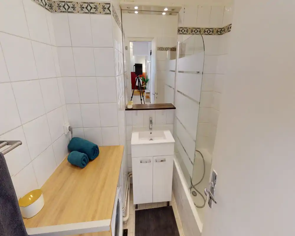 Bagno compatto e ben illuminato con vasca e parete doccia in vetro, piccolo lavabo con mobiletto e piano in legno con asciugamani arrotolati.