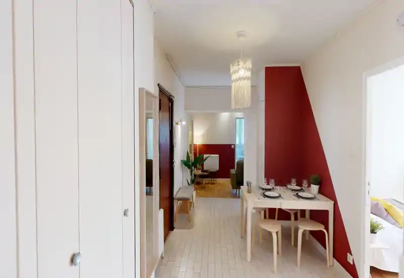 Bright, neatly staged living area seen from the entrance with a small dining table, accent red wall and an open view into a cozy sitting space with warm lighting.