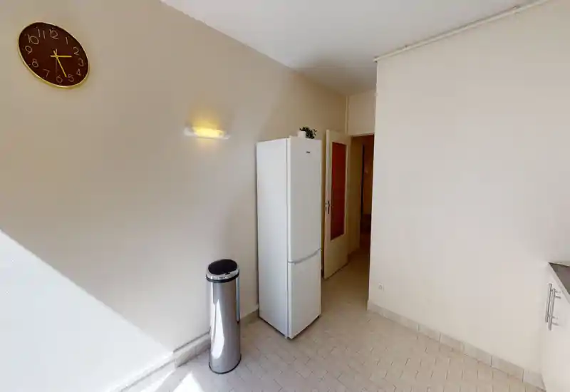 Bright, minimal kitchen corner featuring a freestanding refrigerator, a stainless steel bin and tiled floor — simple, clean layout with natural light.