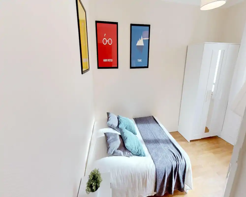 Chambre lumineuse et soignée avec un lit double, coussins texturés et plaid gris ; armoire blanche et sol en bois pour une ambiance minimaliste et apaisante.