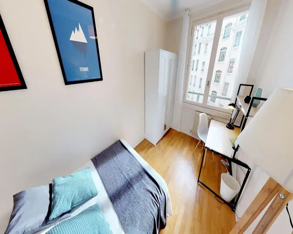 Chambre simple lumineuse et soignée avec lit bien fait, sol en bois, armoire et bureau près d'une grande fenêtre laissant entrer beaucoup de lumière naturelle.