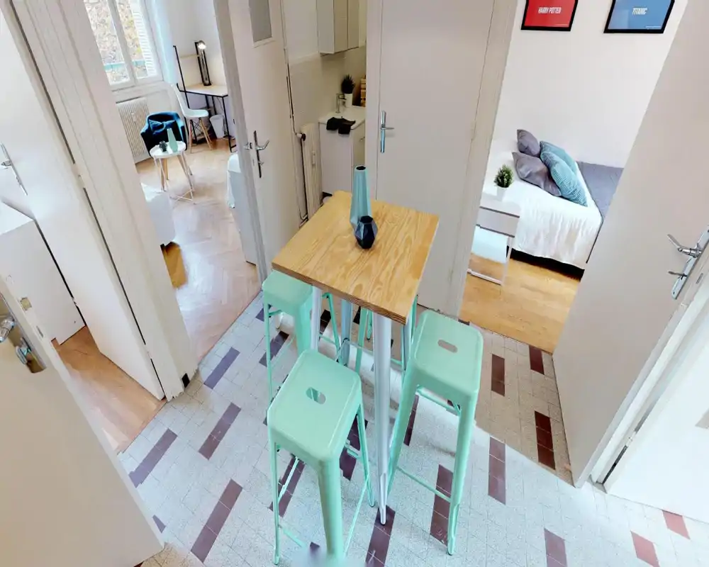 Coin repas/ petit déjeuner compact avec une table haute en bois et quatre tabourets vert menthe sur un carrelage à motif ; espace lumineux et aéré qui communique avec le salon et la chambre.