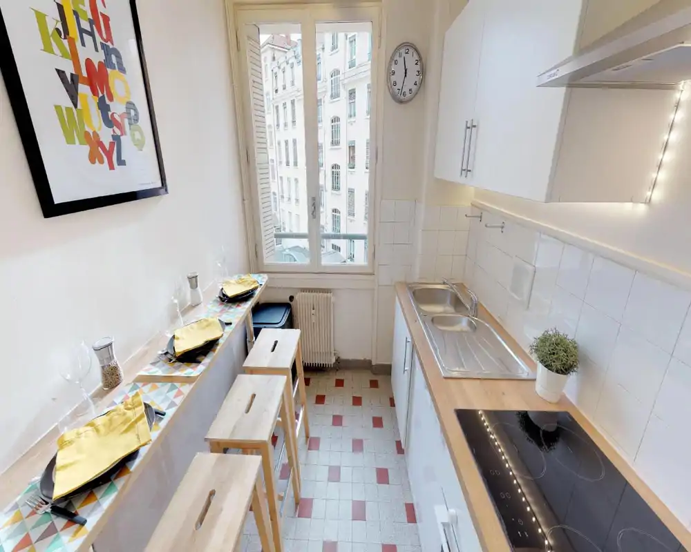 Cuisine compacte et lumineuse avec plan de travail en bois, évier, plaque à induction et un bar étroit avec tabourets — dressée pour les repas et éclairée par une grande fenêtre.