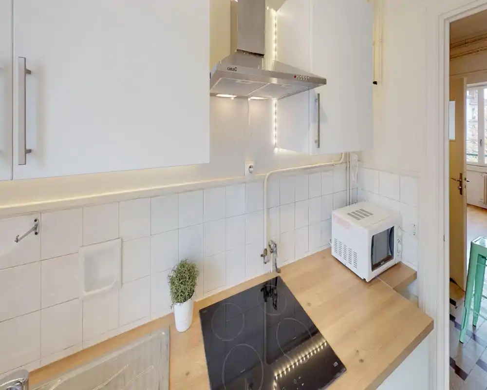 Cuisine compacte et lumineuse avec plaque de cuisson électrique moderne, plan de travail en bois, placards blancs et micro-ondes sur le côté — propre, fonctionnelle et bien éclairée.