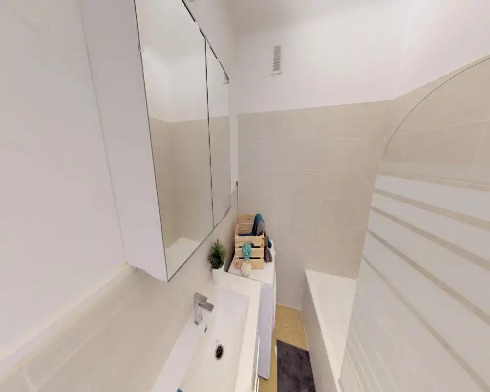 Salle de bain compacte et moderne avec lavabo, armoire miroir et baignoire avec paroi en verre. Carrelage beige neutre et accessoires soignés pour une impression propre et fonctionnelle.