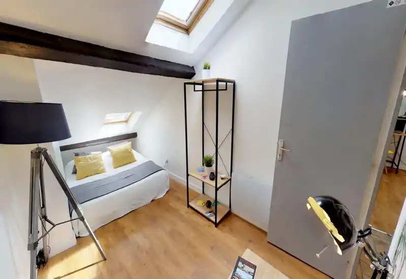 Camera da letto mansardata e accogliente con letto matrimoniale basso sotto lucernari, pavimento in legno caldo, cuscini gialli decorativi e una scaffalatura minimalista — luminosa e invitante.