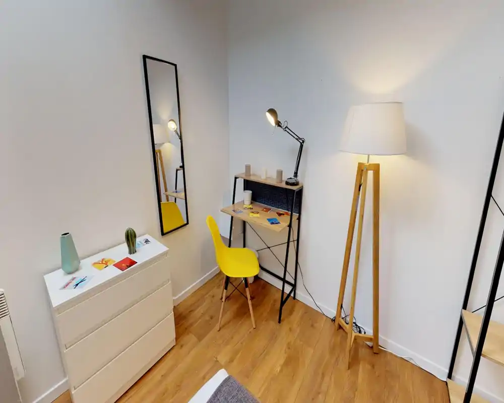 Angolo camera da letto elegante e compatto con una scrivania, sedia gialla vivace e illuminazione calda. Si vede una porzione del letto, pavimento in legno e arredi minimalisti.