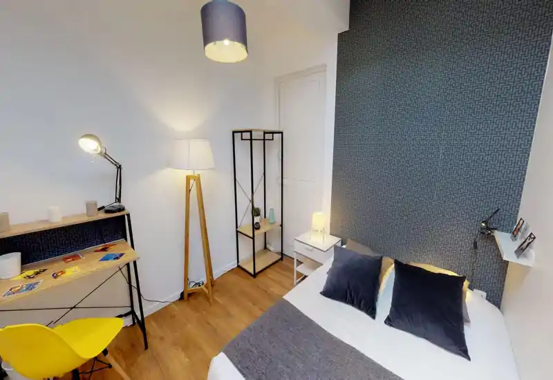Camera da letto moderna e luminosa con letto matrimoniale in lenzuola bianche, cuscini scuri e parete decorativa a motivo. Comodino con lampada, lampada da terra, scaffale e scrivania compatta rendono l’ambiente funzionale e accogliente, perfetto per affitti brevi.