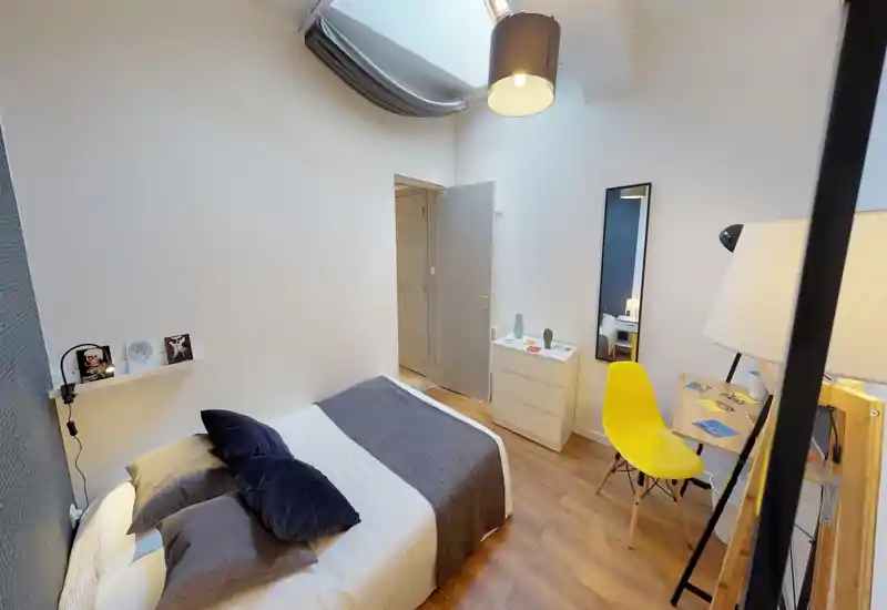 Camera da letto luminosa e moderna con letto ampio, cuscini stratificati e una coperta grigia accogliente. La luce naturale da un lucernario e le lampade calde creano un’atmosfera invitante; una piccola scrivania con sedia gialla e uno specchio completano l’arredamento.