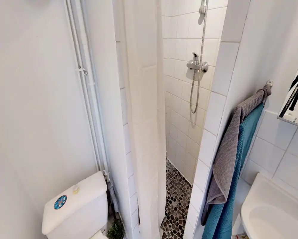 Bagno compatto con doccia a pavimento piastrellata, doccetta, WC e ganci per asciugamani; piastrelle bianche e pavimento a ciottoli donano struttura.