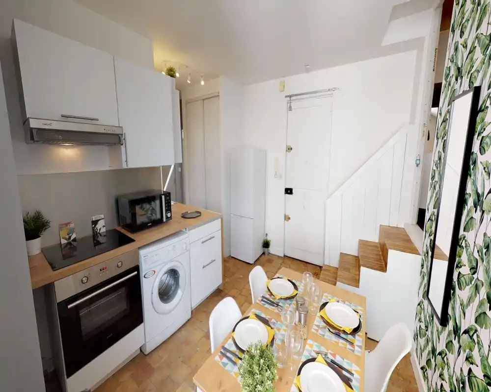 Cucina open-space compatta con mobili bianchi moderni, forno, piano a induzione, lavatrice e tavolo apparecchiato per quattro persone — luminosa, ben curata e pratica per soggiorni brevi.