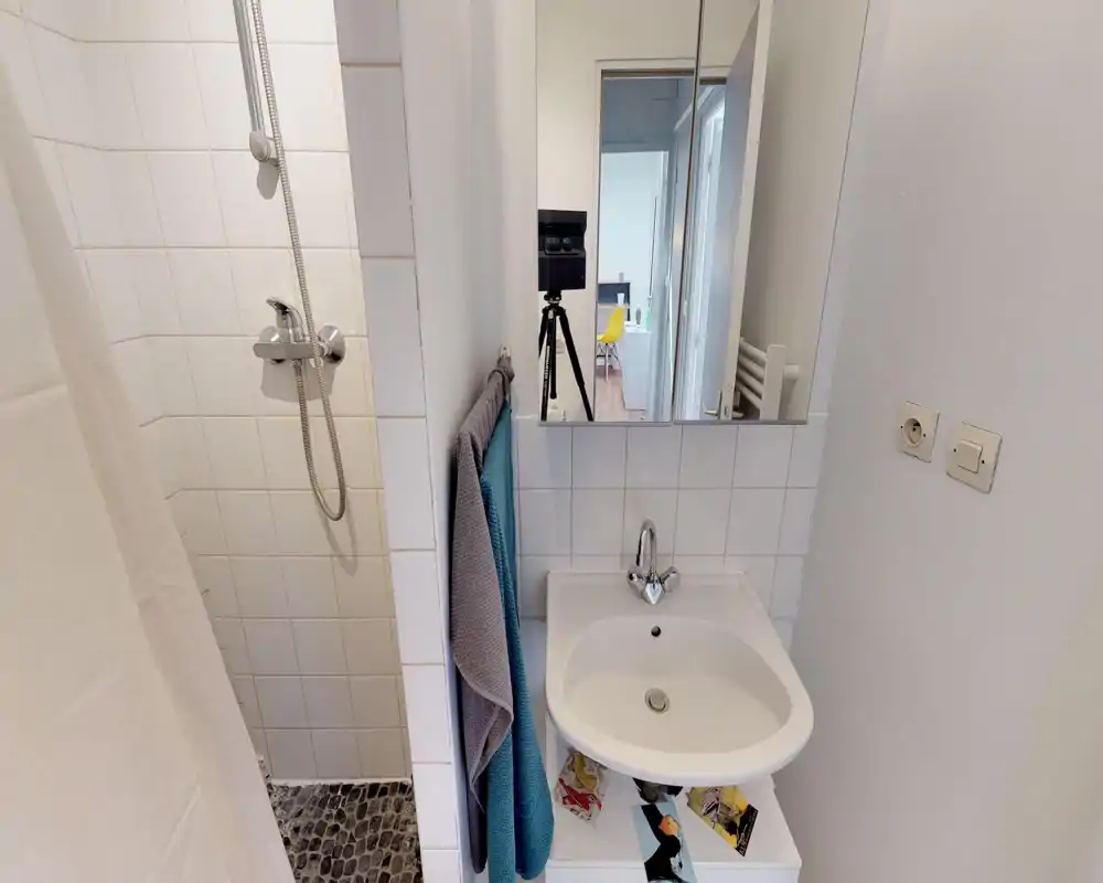 Bagno compatto con doccia piastrellata, lavabo bianco e specchio con mobiletto; luminoso, pulito e funzionale per un piccolo affitto.