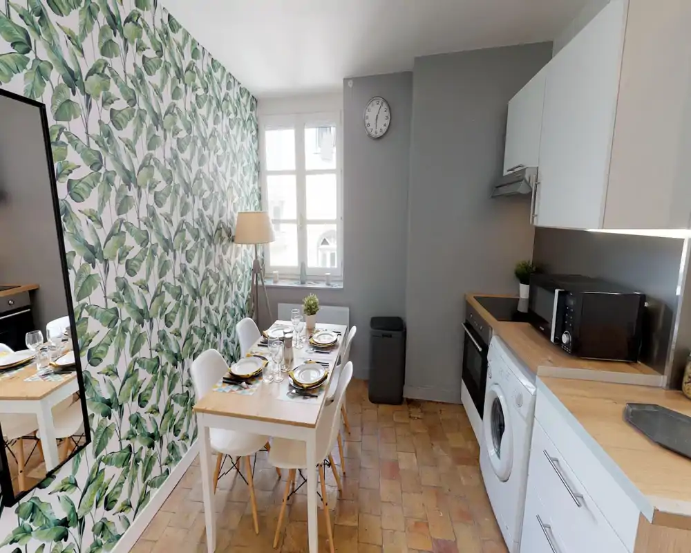 Cucina compatta e di stile con elettrodomestici integrati, piccolo tavolo in legno apparecchiato per quattro e carta da parati a foglie che dona personalità e calore.