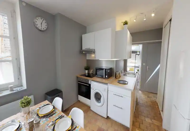Compact modern kitchen with white cabinets, wooden countertops, built-in oven and washing machine; bright, neatly staged with a small dining area visible.