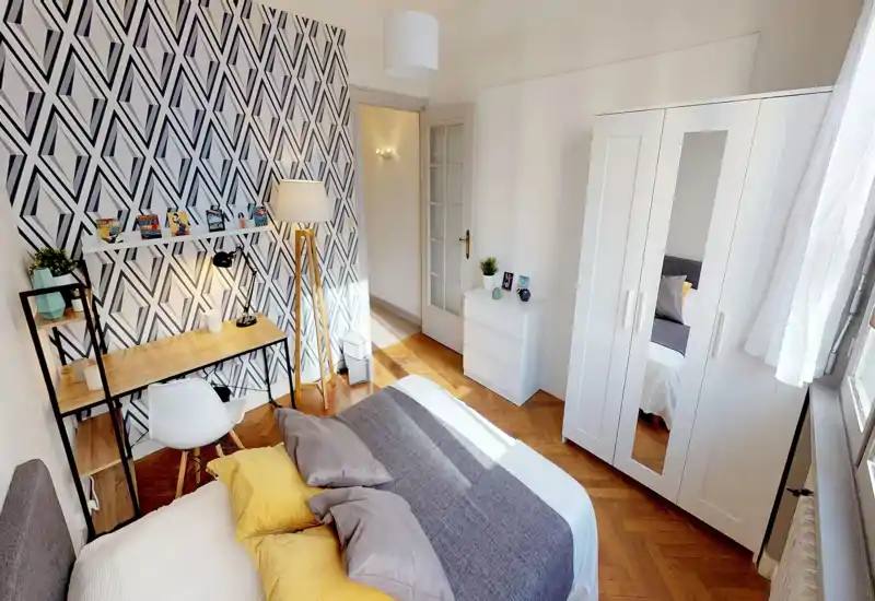 Bright, stylish bedroom with a comfortable double bed dressed in neutral linens and colored cushions, a work desk with shelving, patterned accent wall and a white wardrobe — ideal for a cozy rental.