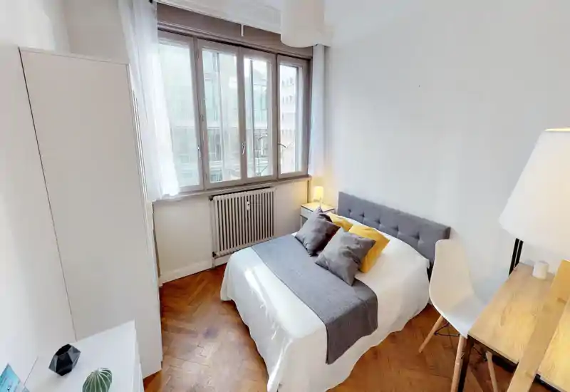 Bright, neatly styled bedroom with a comfortable double bed, soft pillows and a grey headboard; large windows provide natural light and the wooden herringbone floor adds warmth.