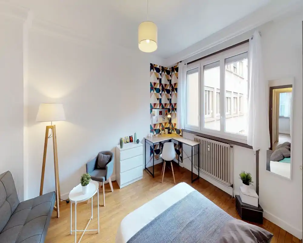 Chambre lumineuse et soignée avec lit au premier plan, coin salon cosy et bureau près de la fenêtre — idéale pour travailler et se détendre.
