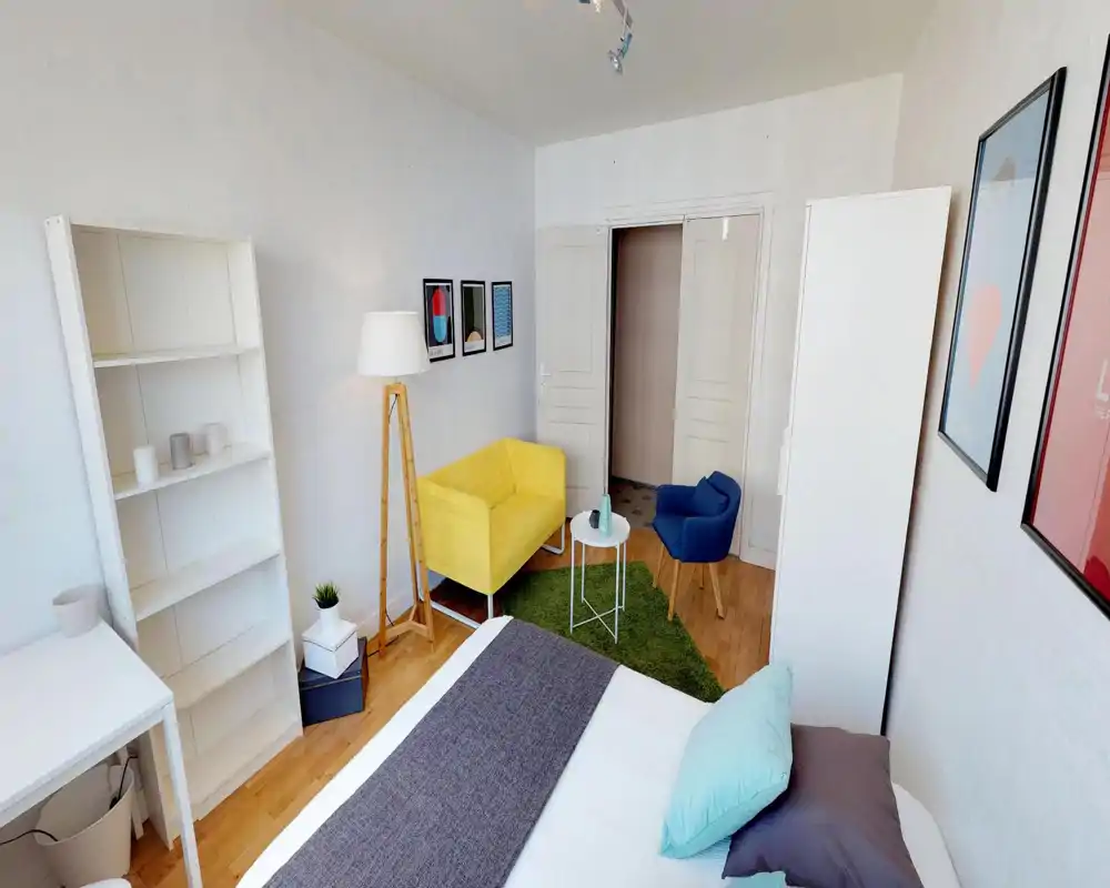Bright, styled bedroom with a visible double bed in the foreground, colorful seating (yellow loveseat and blue armchair), wooden floor and framed artwork — a cozy, well-lit room suitable for a rental listing.