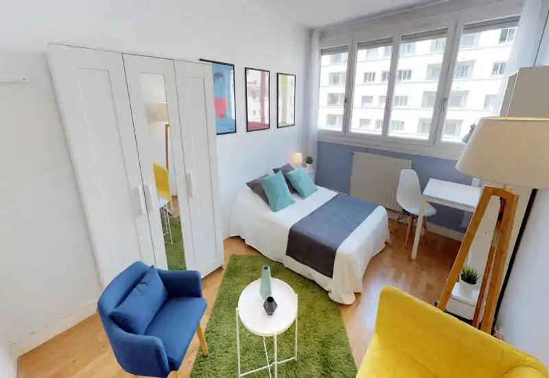 Bright, well-staged bedroom with a double bed, decorative cushions and a green rug; large windows provide abundant natural light and a compact desk by the window for working.