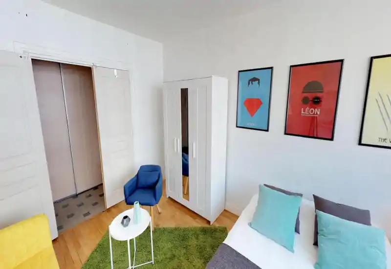 Bright, modern bedroom corner with a visible double bed dressed in white linens and turquoise pillows, a white wardrobe with mirror, a blue armchair and colorful framed posters on the wall. The room feels fresh and inviting with wooden floors and a small green rug.