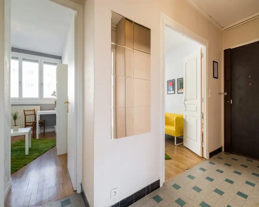 Bright and tidy entrance hallway with tiled floor, front door and views into adjacent rooms including a living space with a yellow sofa.
